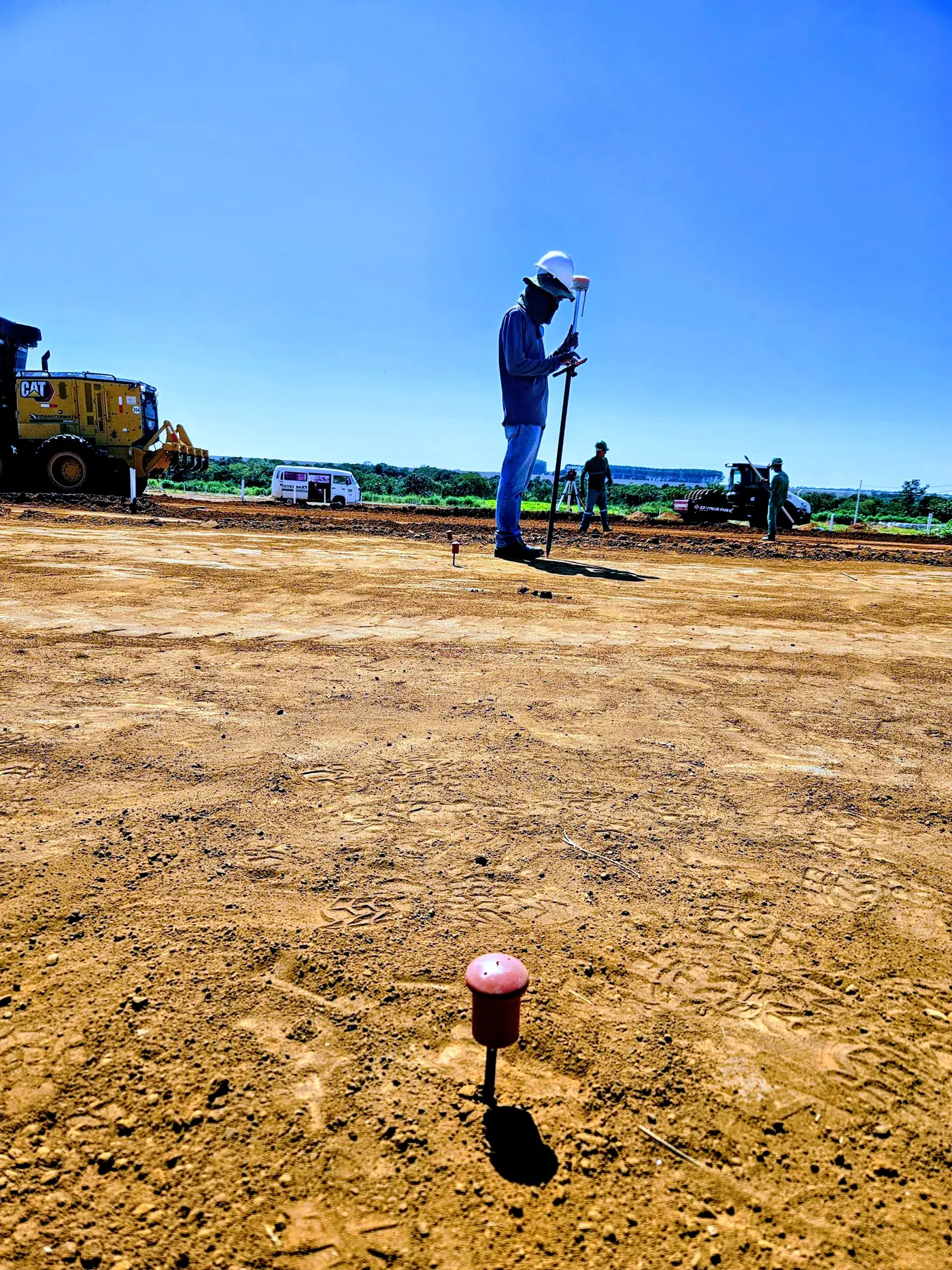 Topógrafo em campo aberto com maquinário de terraplenagem ao fundo